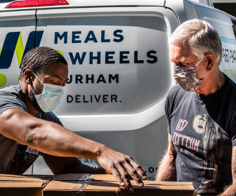 Two man working on wood in front of Meals on Wheels delivery van.