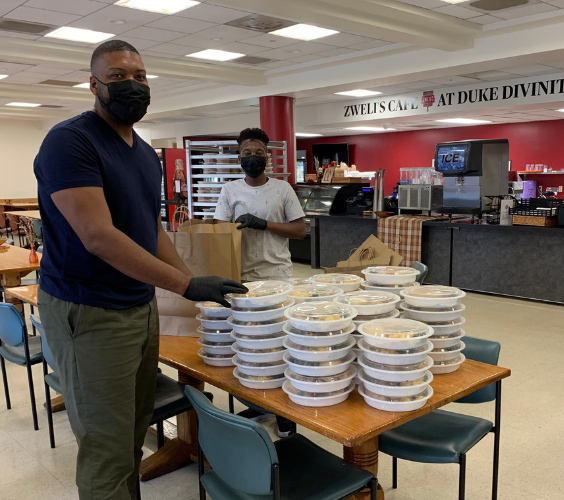 Chef and young chef pose for a pictures as they stack meal trays on a table.