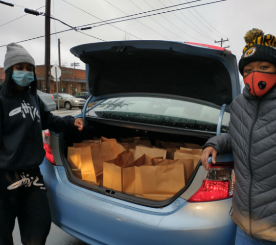 Two Volunteer stands beside the trunk of a sedan with a trunk full of meal bags.
