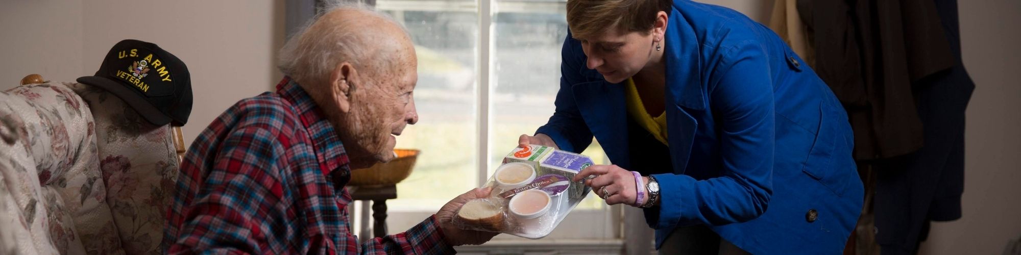 Lady leaning down as she hands older gentleman his food tray on his couch.