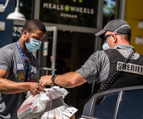 Hand of sheriff deputy handing bags of fruit to a younger volunteer outside the Meals on Wheels Durham office.