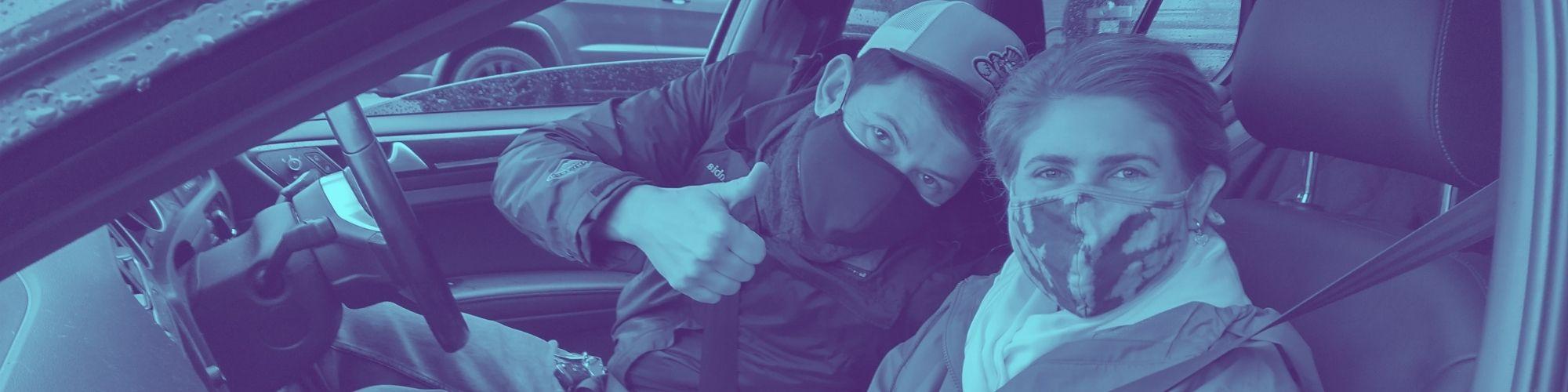 Two volunteers smiling under their mask in the front seat of the car. Male volunteer in the passenger seat is show a thumbs up.