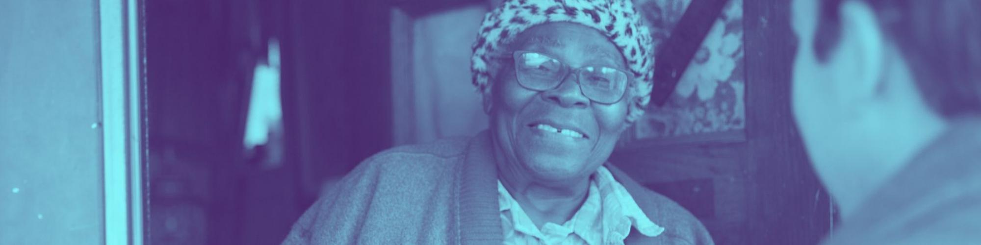 Older lady with fuzzy hat smile at her front door as a volunteer hands her a meal off camera.