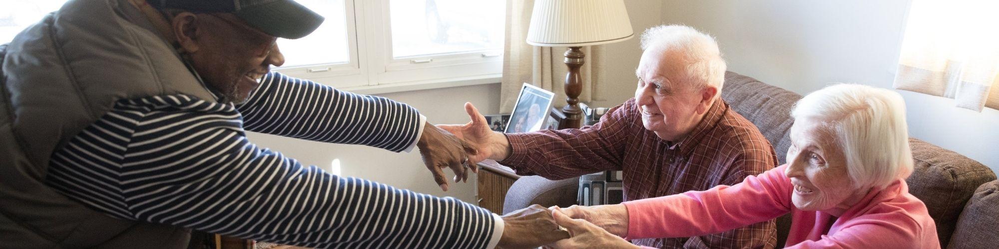 Volunteer reaching out to touch hands of two elderly clients sitting on the count, smiling.