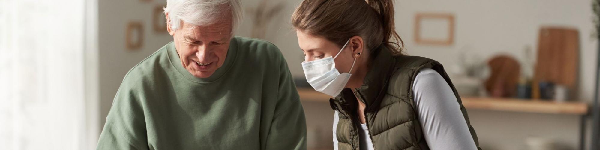 Young lady with face mask leans in to assist older male in kitchen.