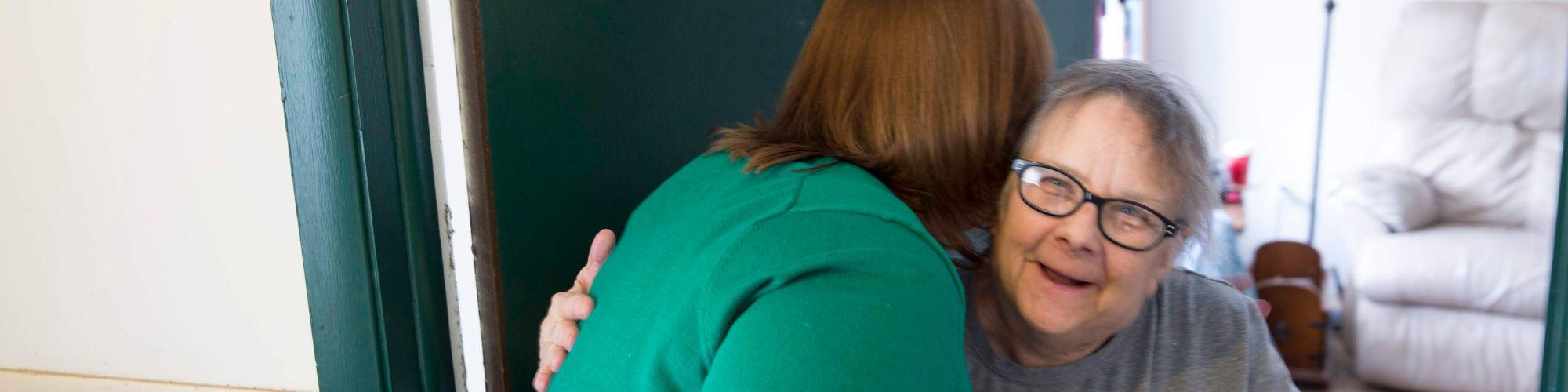 Volunteer in a green long sleeve leans over to hug smiling client the volunteer hands the client her food.