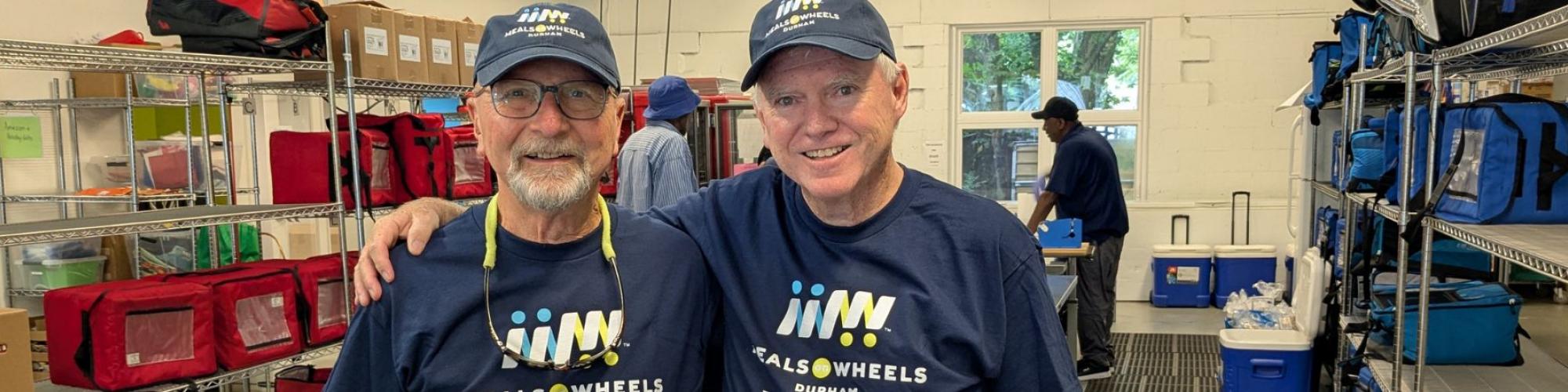 Two Meals on Wheels Durham Volunteer smiling together, both wearing a navy Meals on Wheels Durham hat and t-shirt.